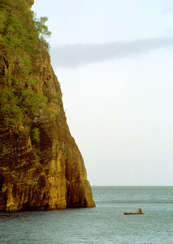 A man paddles his canoe into Lolowei's harbour, sheltered by standing rocks on one side and this massive cliff on the other. 