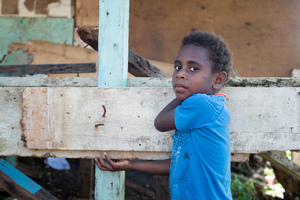 A girl stands beside a half-constructed house made from salvaged materials.
